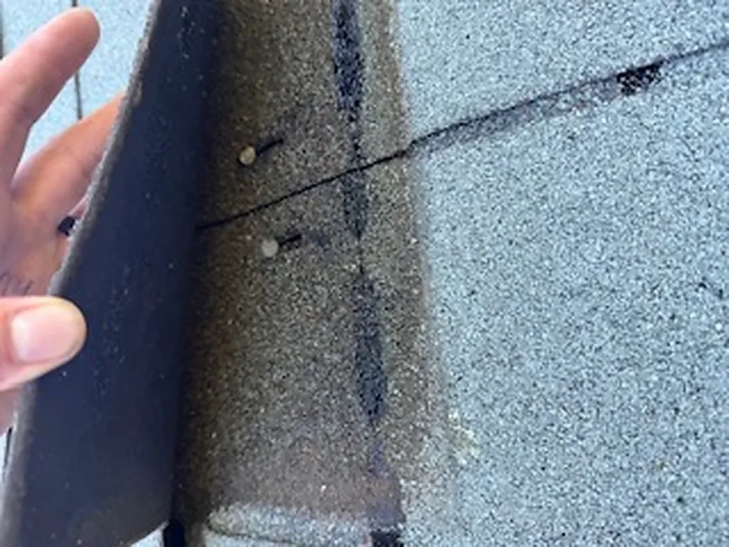 Hand lifting a weathered shingle tab to show the darker covered strip beneath beside the lighter, exposed surface.