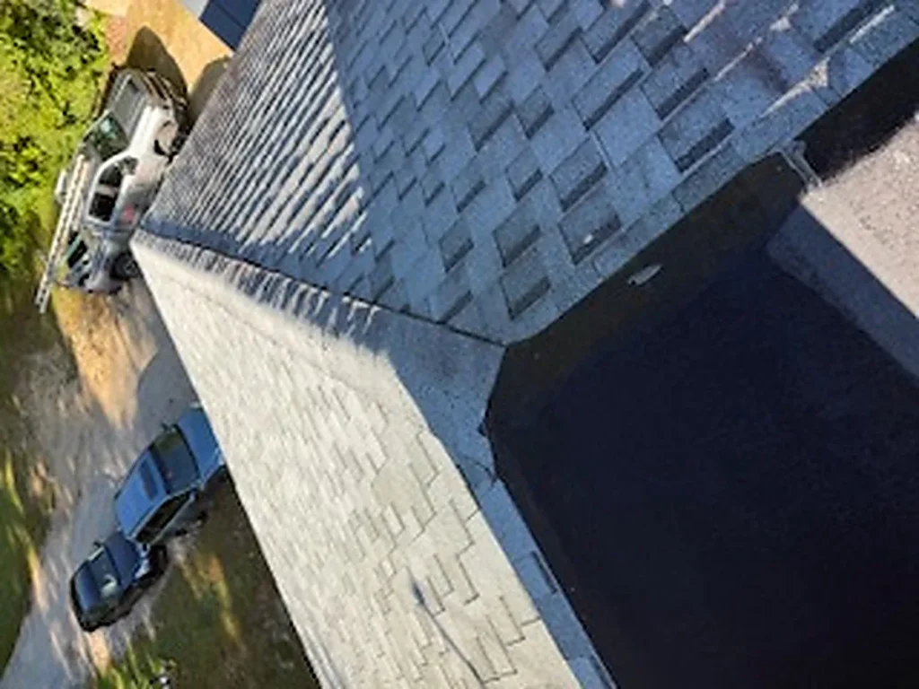 Looking down a pale, weathered asphalt shingle roof from above, with a ladder and vehicles in the driveway below.